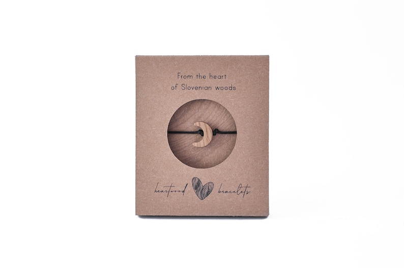 May include: A brown cardboard box with a black string bracelet with a wooden crescent moon charm. The box has the text "From the heart of Slovenian woods" and "heartwood bracelets".