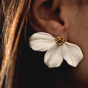 Op de afbeelding: Een witte bloemvormige oorbel met een goudkleurig middenstuk. De oorbel wordt op een oor gedragen, met het haar en het oor van de persoon zichtbaar. De oorbel heeft drie bloemblaadjes en een getextureerd midden.