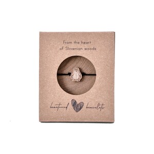May include: A brown cardboard box with the text "From the heart of Slovenian woods" and "heartwood bracelets". A wooden penguin charm is displayed on a black string bracelet inside the box.