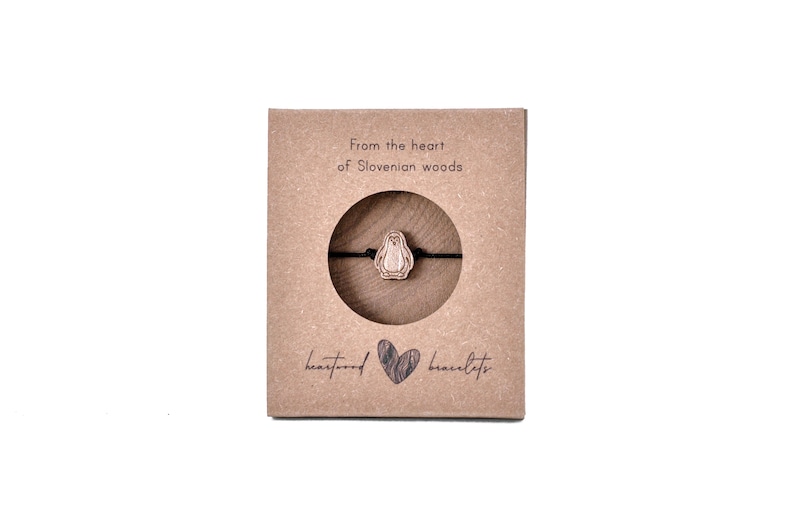 May include: A brown cardboard box with the text "From the heart of Slovenian woods" and "heartwood bracelets". A wooden penguin charm is displayed on a black string bracelet inside the box.