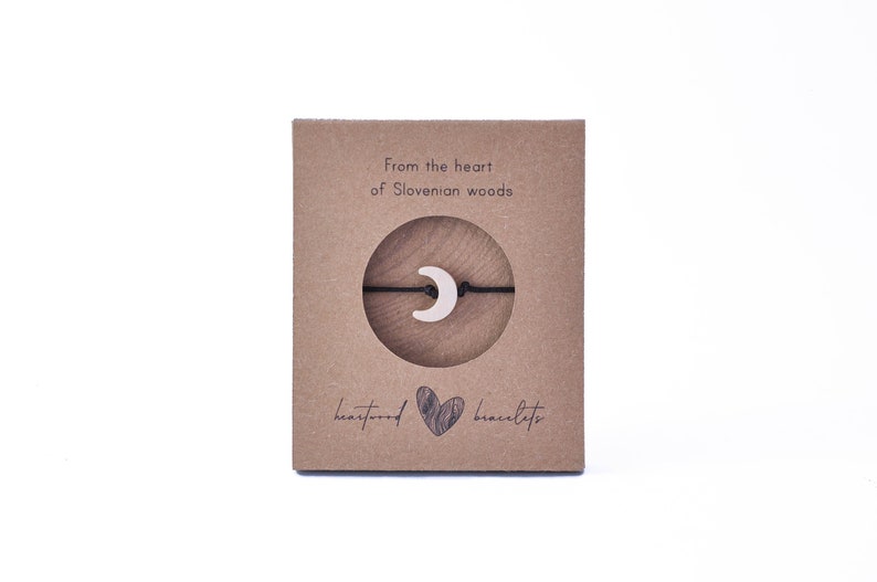 May include: A brown cardboard box with the text "From the heart of Slovenian woods" and "heartwood bracelets". A black string bracelet with a white crescent moon charm is displayed on the box.