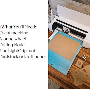 May include: A white Cricut cutting machine with a blue cutting mat and a brown piece of cardstock on top. The cardstock has been cut into a shape with a scoring wheel. The image includes the text "What You'll Need: - Cricut machine - Scoring wheel - Cutting blade - Blue LightGrip mat - Cardstock or kraft paper"