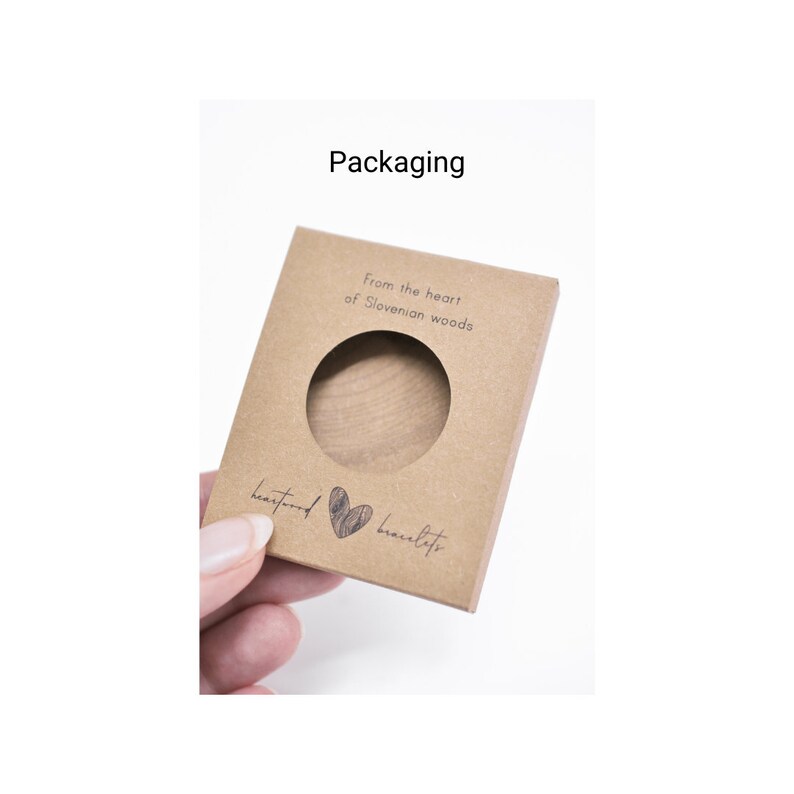 May include: A brown cardboard box with a cut-out window, showcasing a wooden bracelet. The box has the text "From the heart of Slovenian woods" and "heartwood bracelets" printed on it.