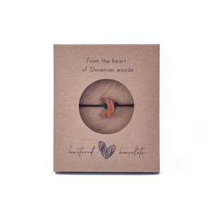 May include: A brown cardboard box with the text "from the heart of Slovenian woods" and "heartwood bracelets". The box contains a wooden crescent moon charm on a black string.