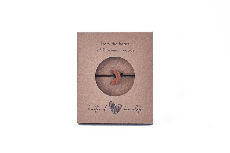 May include: A brown cardboard box with the text "from the heart of Slovenian woods" and "heartwood bracelets". The box contains a wooden crescent moon charm on a black string.