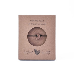 May include: A brown cardboard box with the text "From the heart of Slovenian woods" and "heartwood bracelets". The box contains a wooden crescent moon charm on a black string.