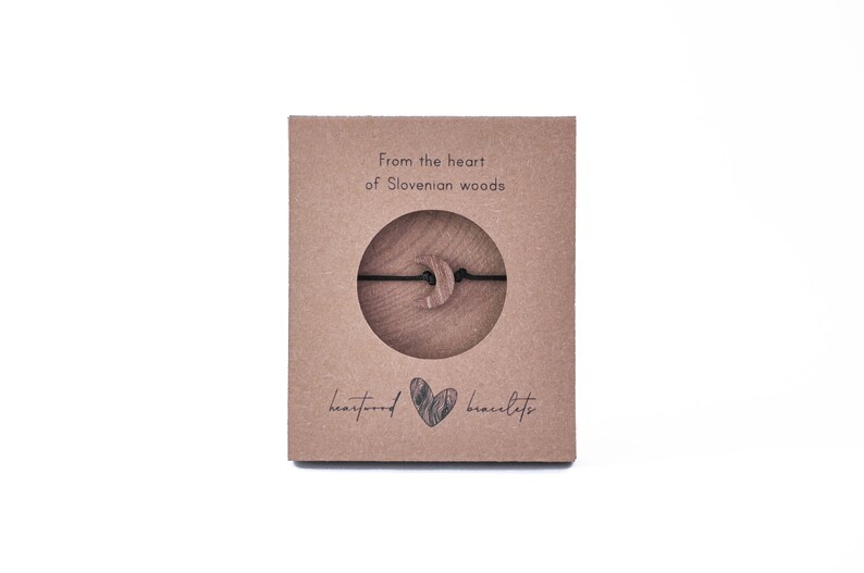 May include: A brown cardboard box with the text "From the heart of Slovenian woods" and "heartwood bracelets". The box contains a wooden crescent moon charm on a black string.