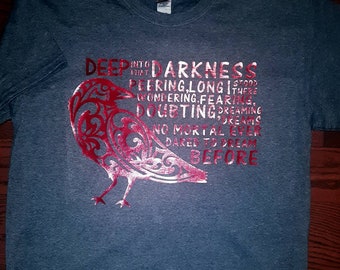 Op de afbeelding: Een donkergrijs T-shirt met een rood en wit gestileerd vogelontwerp. Het shirt heeft de tekst "DEEP INTO THAT DARKNESS STOOD PEERING LONG THERE WONDERING FEARING, DOUBTING DREAMING, NO MORTAL EVER DARED TO DREAM BEFORE" erop gedrukt.