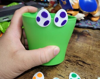 以下が含まれることがあります： 異なる色の斑点のある白いプラスチック製の卵が4つ。2つの卵は紫色の斑点があり、1つはオレンジ色の斑点があり、1つは緑色の斑点があります。