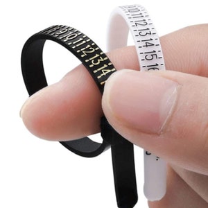 May include: Black and white ring sizers with numbered markings. The black sizer has gold numbers, and the white sizer has black numbers. The sizers are flexible and used to measure ring sizes. The image shows the sizers being held by a person.