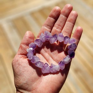 May include: A bracelet made of raw, uncut amethyst stones. The bracelet is a light purple color and is held in an open hand. The stones are irregularly shaped and have a natural, organic appearance. The background is a light brown color.