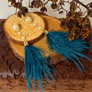 May include: Gold-toned earrings with faux pearl studs and dangling teal feathers. The earrings are connected by a delicate chain of small pearls. The feathers are a vibrant teal color, adding a pop of color to the design.
