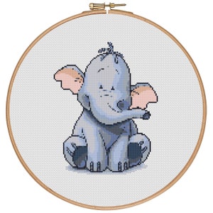 Puede incluir: Un bordado de punto de cruz de un elefante bebé en un aro de madera. El elefante es azul claro con orejas rosas y está sentado. El bordado está sobre un fondo blanco.