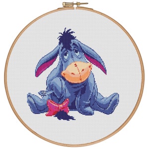 Pode incluir: Um padrão de ponto cruz bordado de um burro triste com um laço rosa. O burro está sentado com as pernas cruzadas e a cabeça inclinada para baixo.