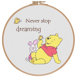 Può includere: Un cerchio da ricamo bianco con uno sfondo grigio chiaro che presenta Winnie the Pooh e Piglet. Winnie the Pooh tiene una farfalla e Piglet è seduto accanto a lui. Il testo "Never stop dreaming" è scritto in marrone sopra di loro.