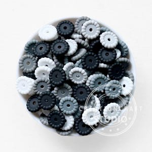 Può includere: Una ciotola bianca piena di perline in silicone a forma di fiore, nei toni del bianco, grigio e nero. Queste perline sono probabilmente destinate al fai-da-te o alla creazione di gioielli. L'immagine è ben illuminata e a fuoco.