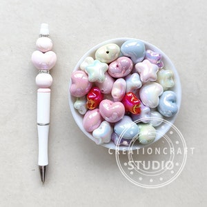 Puede incluir: Un bol lleno de cuentas iridiscentes de color rosa pastel, azul y blanco en forma de corazón y estrella. Las cuentas están en un bol blanco con un logotipo en blanco y negro que dice "Creationcraft Studio".