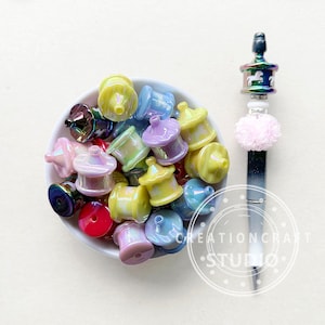 Puede incluir: Un bol lleno de cuentas de vidrio iridiscente de colores con forma de miniaturas de atracciones de feria. Las cuentas son de color rosa, morado, amarillo y azul.