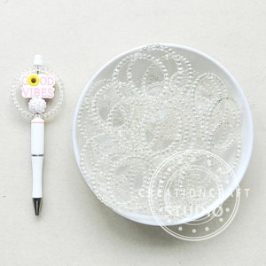 以下が含まれることがあります： 透明なガラスビーズで満たされた白いボウル。ボウル横に、「Good Vibes」と書かれたピンクと黄色の花のチャームが付いた白いペンがあります。