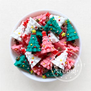 Puede incluir: Un bol lleno de adornos con forma de árbol de Navidad de colores. Los adornos están hechos de resina y tienen un acabado brillante. Están decorados con espolvoreados blancos, rosas, verdes y rojos.