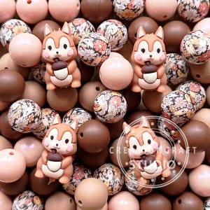 May include: A collection of brown, pink, and white silicone beads with a floral pattern and a squirrel design. The beads are strung together to create a necklace or bracelet.