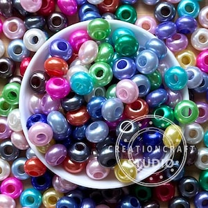 Puede incluir: Una colección de cuentas coloridas y nacaradas en una variedad de tonos, incluyendo rosa, verde, azul y morado. Las cuentas están dispersas alrededor de un cuenco blanco con el texto "CREATION CRAFT STUDIO" en un logotipo circular.