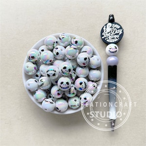 May include: A white bowl filled with iridescent white beads featuring a black Jack Skellington face design. The beads are in a white bowl on a white surface. A pen with a black and white design and a round black charm with the text "Have the Day You Deserve" is next to the bowl.