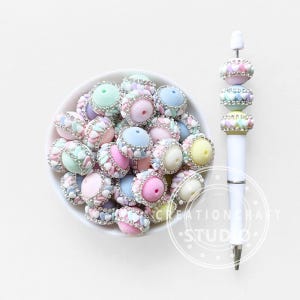 May include: A white bowl filled with pastel-colored beads with rhinestone accents. A white pen with decorative beads in matching colors sits to the right. The beads are in shades of pink, blue, yellow, and green.