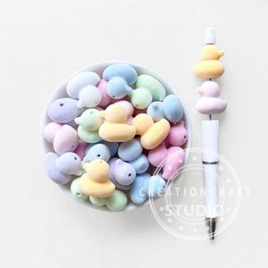 Puede incluir: Un cuenco blanco lleno de caramelos con forma de pato en colores pastel. A la derecha, un bolígrafo blanco con una pila de adornos con forma de pato en amarillo, rosa y naranja. Los caramelos y el bolígrafo están sobre una superficie blanca.