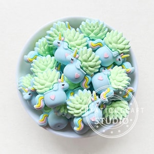 May include: A bowl filled with light blue and green unicorn charms. Each charm features a unicorn with a green succulent on its back. The charms are made of silicone and are perfect for adding a touch of whimsy to any project.