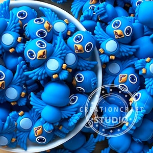 Può includere: Una collezione di abbellimenti blu a forma di uccello con occhi bianchi e becchi arancioni. Gli uccelli hanno le ali spiegate e sono ammucchiati in una ciotola bianca. L'immagine include il testo "CREATIONCRAFT STUDIO".