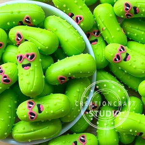 Peut inclure: Collection de breloques en forme de cornichons vert vif, certaines avec des lunettes de soleil roses en forme de cœur et des visages souriants. Les breloques sont éparpillées, certaines dans un bol blanc. L'image comprend le texte "CREATIONCRAFT STUDIO".