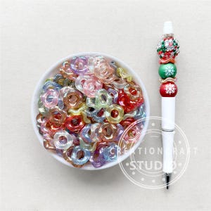 May include: A bowl filled with iridescent rainbow colored plastic donut shaped beads. The beads are perfect for jewelry making or crafting.
