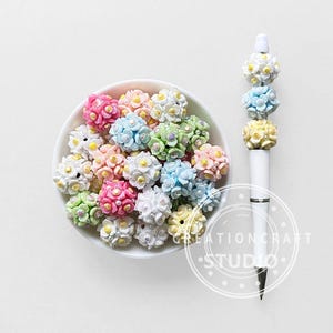 May include: A white bowl filled with colorful, flower-shaped beads in shades of pink, blue, green, and yellow. A white pen with flower-shaped embellishments in matching colors is also present. The image is set against a white background.