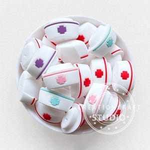 Pode incluir: Uma tigela cheia de encantos de chapéu de enfermeira de silicone branco. Os encantos têm acentos vermelhos, rosados, roxos e azuis. Os encantos são decorados com uma cruz vermelha.