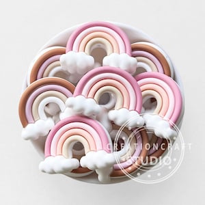 Puede incluir: Una colección de decoraciones en forma de arcoíris en colores pastel con detalles de nubes blancas. Los arcoíris presentan tonos de rosa, melocotón y marrón. La imagen incluye el texto "CREATIONCRAFT STUDIO" en un diseño circular.