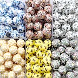 May include: Six containers of colourful silicone beads. The beads are arranged in rows and are all different patterns. The patterns include blue and white floral, brown and white floral, yellow with black bees, white with black butterflies, yellow and white chequered, and purple and white floral.