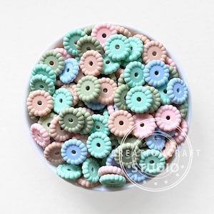 Puede incluir: Un cuenco lleno de cuentas de silicona de colores pastel. Las cuentas tienen forma de flor con un agujero en el centro. Los colores incluyen rosa, azul, verde y melocotón. Las cuentas son probablemente para manualidades o para hacer joyas.