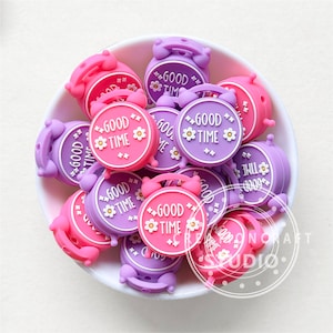 Könnte beinhalten: Eine Schüssel gefüllt mit lila und rosa Silikon-Wecker-Anhängern. Die Anhänger haben weißen Text mit der Aufschrift "Good Time" und einem kleinen Blumendesign.
