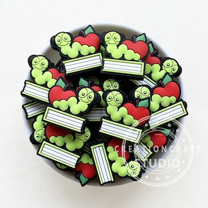 May include: A bowl filled with green and red bookworm charms. Each charm features a green worm with a red apple and a white book with black lines. The charms are in a white bowl with a black and white polka dot pattern. The text "Creationcraft Studio" is visible in the bottom right corner of the image.