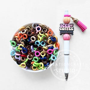 May include: A white bowl filled with colorful jump rings sits next to a white pen with a pink tassel and the words "ICED COFFEE junkie". The pen has pink and orange decorative beads.