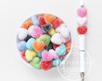 Puede incluir: Un cuenco blanco lleno de caramelos coloridos en forma de corazón. A la derecha, un bolígrafo blanco con una serie de decoraciones en forma de corazón en rosa, rosa claro y rojo. Los caramelos y el bolígrafo están sobre un fondo blanco.