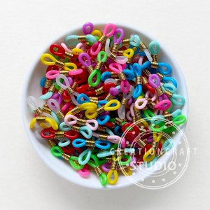 May include: A bowl filled with colorful plastic eyeglass chain connectors. The connectors are in various colors, including red, blue, green, yellow, pink, and clear. Each connector has a gold metal spring.