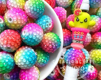 以下が含まれることがあります： 虹色の光沢のある丸いビーズとサボテンのチャームのクローズアップ。ビーズはプラスチック製で、表面にテクスチャがあります。サボテンのチャームはプラスチック製で、緑色のボディにピンクの花、白地にピンクと白のパターンの鉢が付いています。
