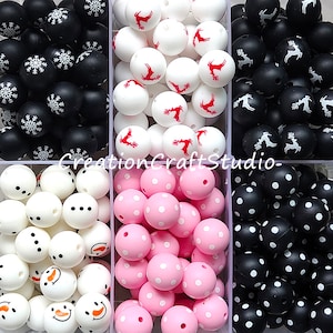 Peut inclure: Assortiment de perles en silicone de différentes couleurs et motifs. Les perles sont disposées en quatre rangées. La première rangée contient des perles noires avec des flocons de neige blancs. La deuxième rangée contient des perles blanches avec des rennes rouges. La troisième rangée contient des perles blanches avec des points noirs. La quatrième rangée contient des perles noires avec des points blancs. CreationCraftStudio-