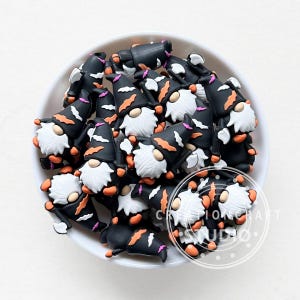 Puede incluir: Un bol lleno de abalorios de gnomo de Halloween negros, naranjas y blancos. Los gnomos llevan sombreros de bruja con murciélagos y un acento rosa.