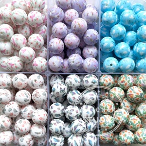 以下が含まれることがあります： さまざまなパターンが施されたシリコンビーズが6つの容器に入っています。ビーズは白で、花柄、蝶、幾何学模様など、さまざまな色のパターンが施されています。