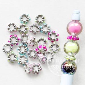 May include: Silver-coloured flower-shaped beads with pink, blue, and green pearl accents. A pen with a light blue top and a series of colourful glass orbs is also visible.