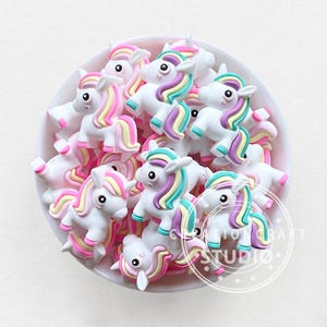 Puede incluir: Un bol lleno de encantos de unicornio coloridos. Los encantos están hechos de arcilla polimérica blanca y tienen acentos rosados, amarillos y azules. Los encantos son perfectos para agregar un toque caprichoso a las manualidades y las joyas.