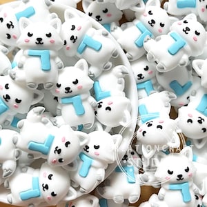 May include: A collection of small white fox-shaped resin charms, each featuring a light blue scarf, black eyes, a black nose, and pink cheeks. The fox figures are arranged in a circular white bowl, with some overlapping and others scattered.